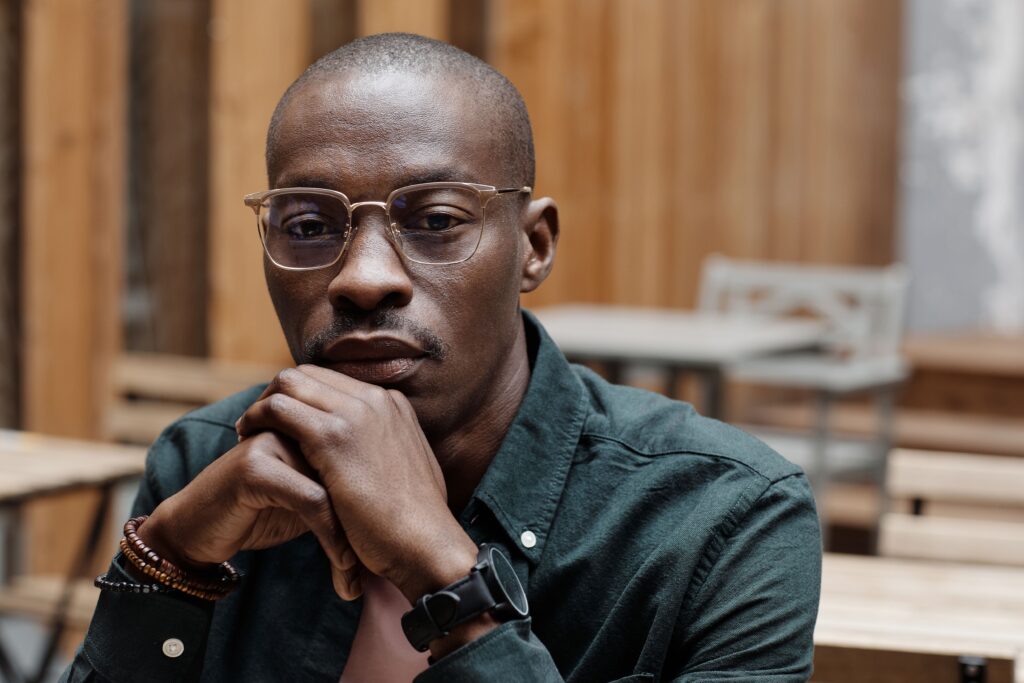 A contemplative man with eyeglasses sits indoors, hands clasped.