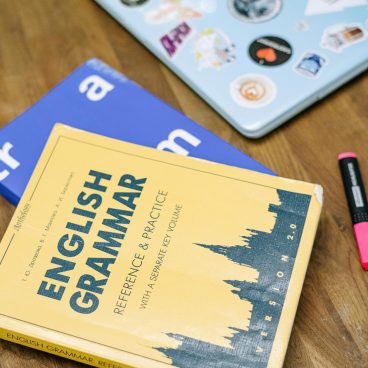 pexels photo 5676740 5676740 High-angle view of an English grammar book and a laptop with stickers on a wooden desk.